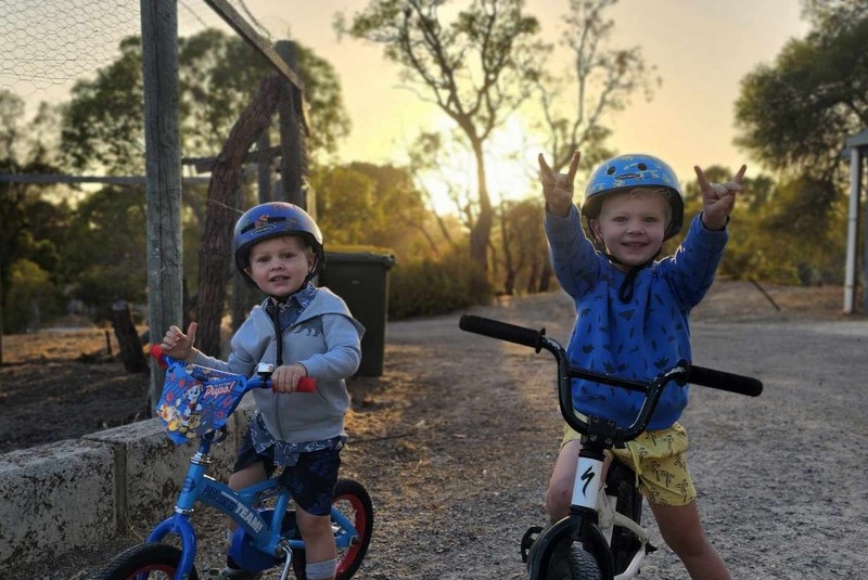 Two kinder-age brothers with blond hair ride on their bikes during sunset. Both are wearing helmets and one is gesturing to the camera.